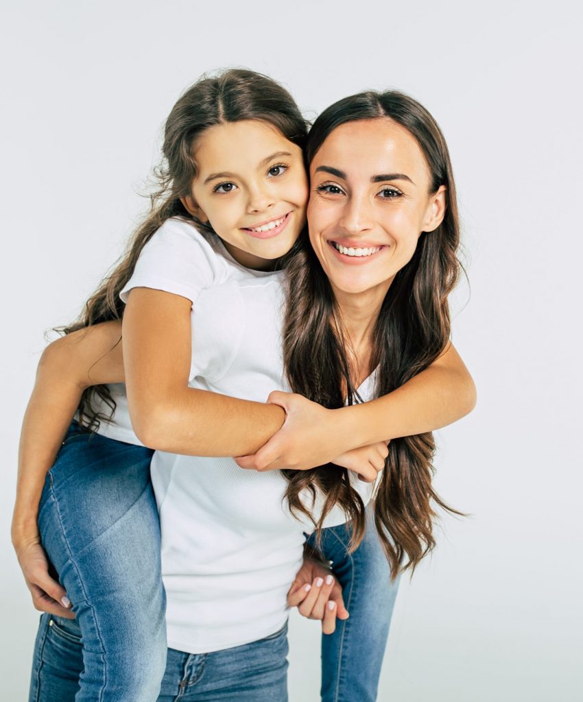 Beautiful young brunette mother holds on back her cute little daughter in white T-shirts isolated in studio